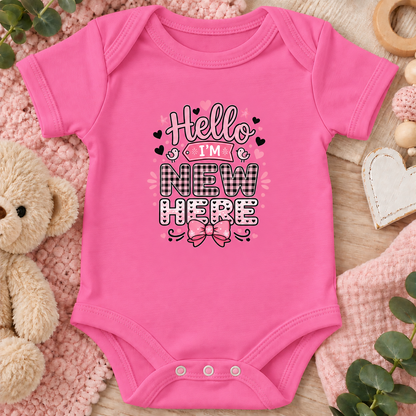 A raspberry colored baby bodysuit with a 'Hello I'M NEW HERE' graphic printed on the chest, styled in a flat-lay with a teddy bear, knit blankets, and wooden toys.