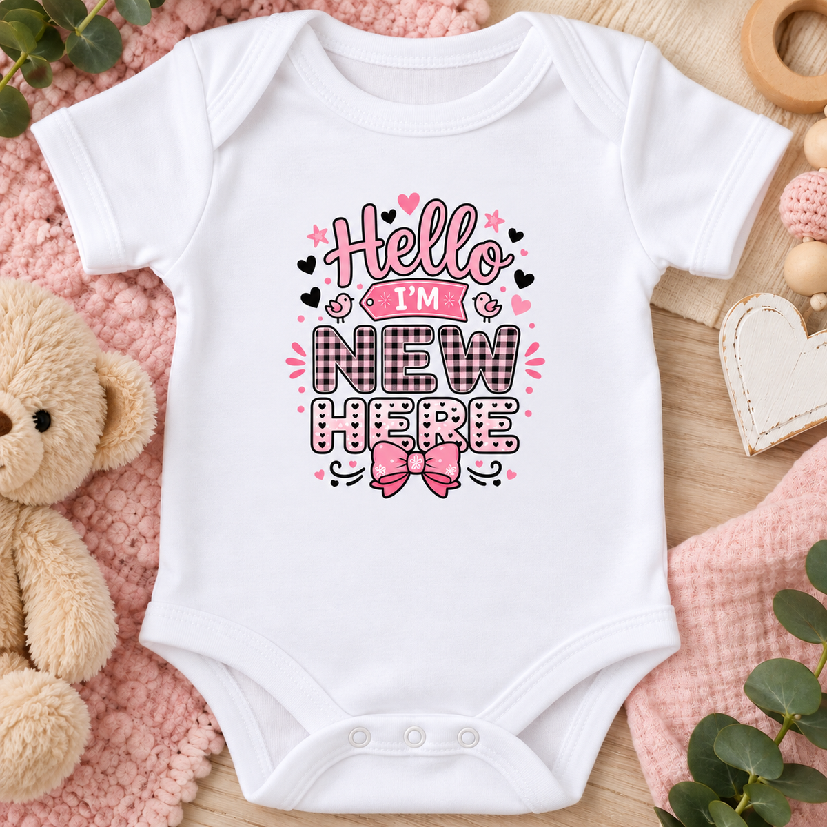 A white short-sleeve baby bodysuit laid flat on a pink textured blanket with a teddy bear, wooden heart, and green leaves around it. The bodysuit has a colorful graphic on the chest that reads “Hello I’m New Here” in pink, black, and white lettering with small heart and star accents.