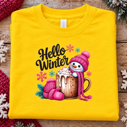 A folded bright yellow T-shirt laid flat on a rustic wooden surface, featuring a colorful winter graphic with the words 'Hello Winter,' a snowman in a pink hat peeking out of a hot cocoa mug, pink mittens, snowflakes, and pine branches.