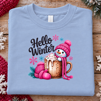 A folded light blue crewneck t-shirt with a 'Hello Winter' graphic print, displayed on a rustic wooden background with holiday decorations.