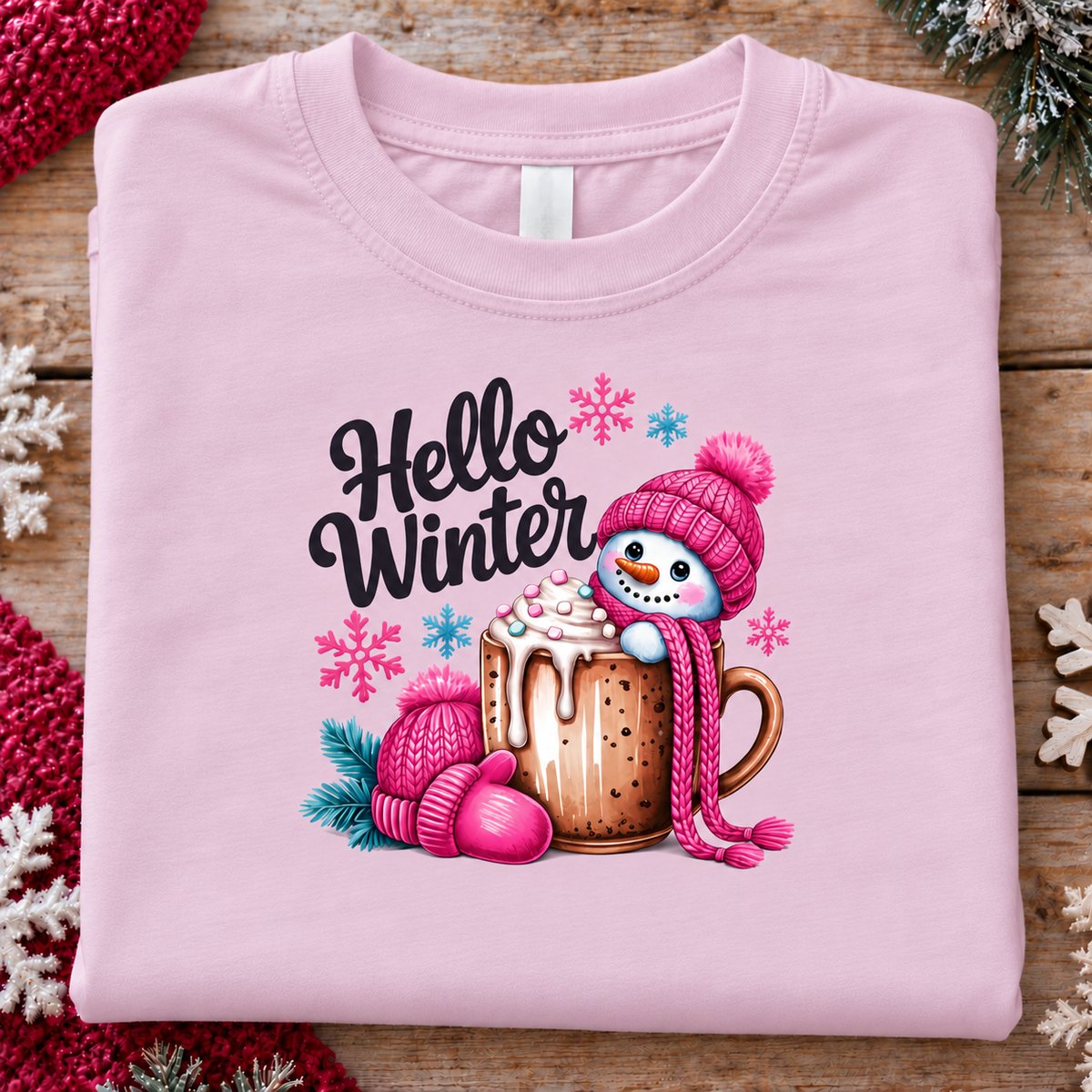 A folded light pink crewneck t-shirt is displayed on a rustic wooden surface, decorated with holiday-themed props like pine branches and knitted scarves. The shirt features a colorful graphic print of a snowman wearing a pink knit hat and scarf, sitting on a mug of hot cocoa with whipped cream, surrounded by pink and blue snowflakes, with the text 'Hello Winter' written in black script.