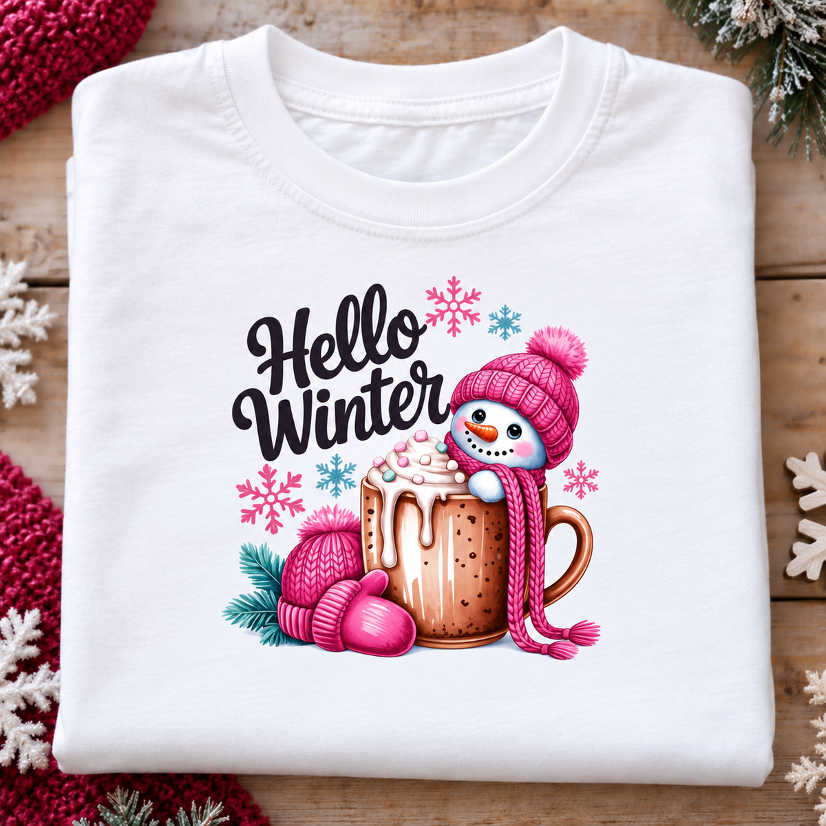 A folded white crewneck t-shirt with a colorful 'Hello Winter' graphic, displayed on a rustic wooden background with festive red and white knit accents.