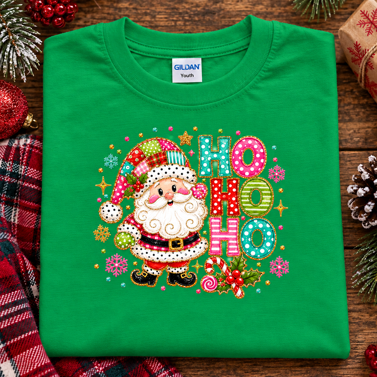 A folded irish green Gildan Youth t-shirt featuring a colorful embroidered Christmas design with a Santa Claus character and 'HO HO HO' lettering, styled on a rustic wooden background with holiday decorations.