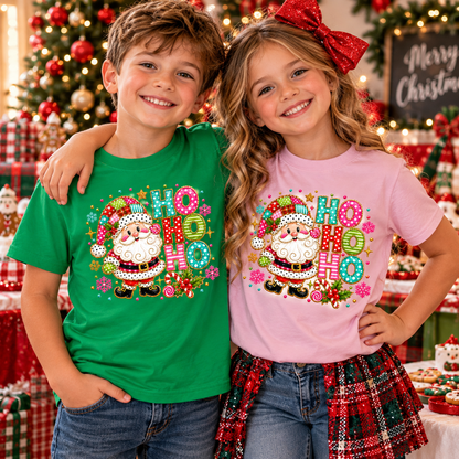 A boy and a girl standing together in a festive Christmas room, wearing matching graphic t-shirts. The boy wears a green t-shirt and the girl wears a pink t-shirt, both featuring a colorful Santa Claus and 'Ho Ho Ho' design.