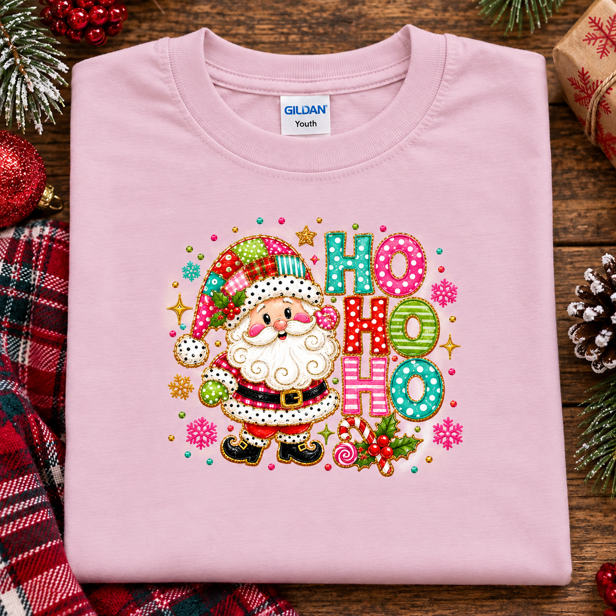 A folded light pink Gildan Youth t-shirt with a colorful Christmas graphic of Santa Claus and 'HO HO HO' text, styled on a rustic wooden background with holiday decorations.