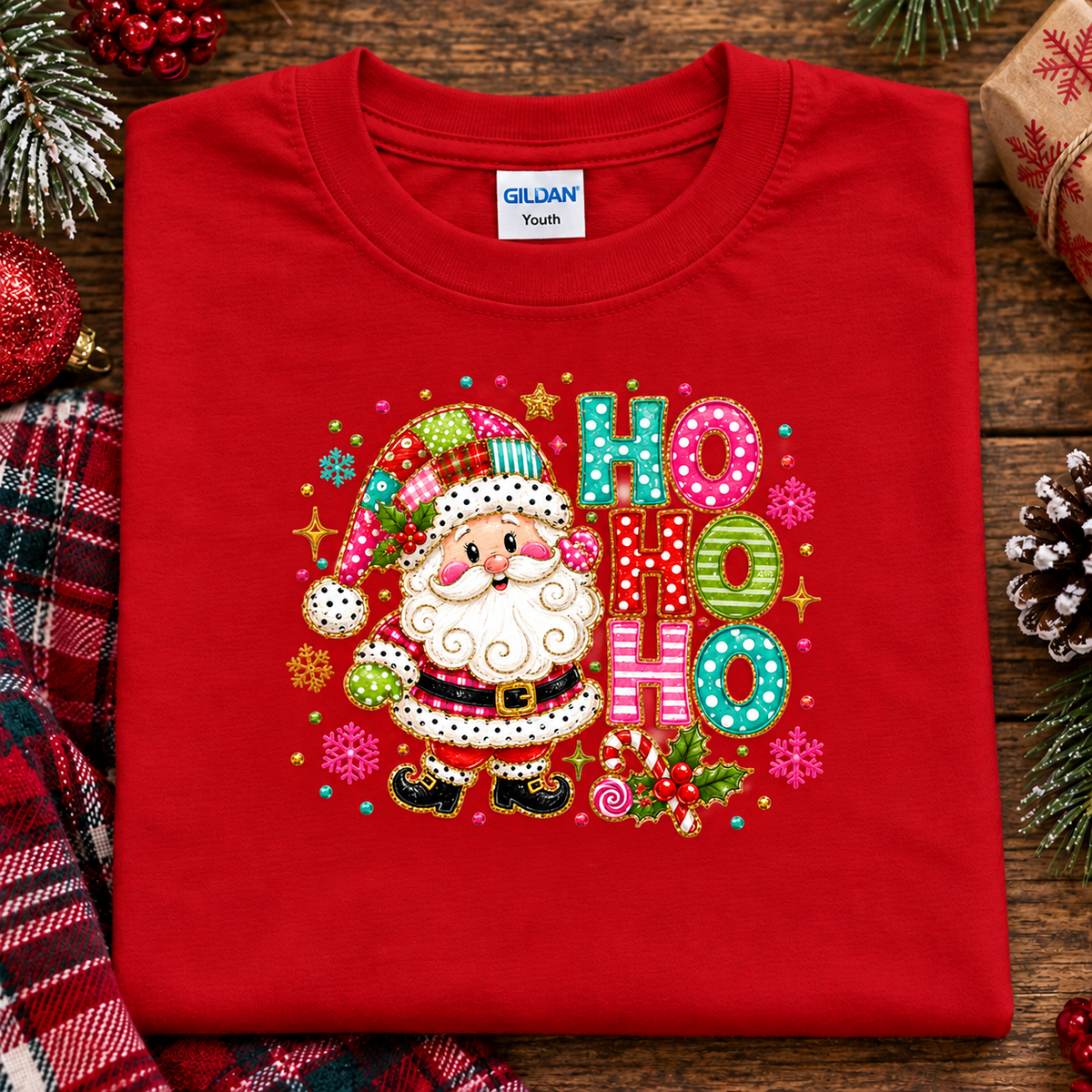 A folded red Gildan Youth t-shirt with a colorful 'Ho Ho Ho' Christmas graphic featuring Santa Claus, displayed on a rustic wooden background with holiday decorations.