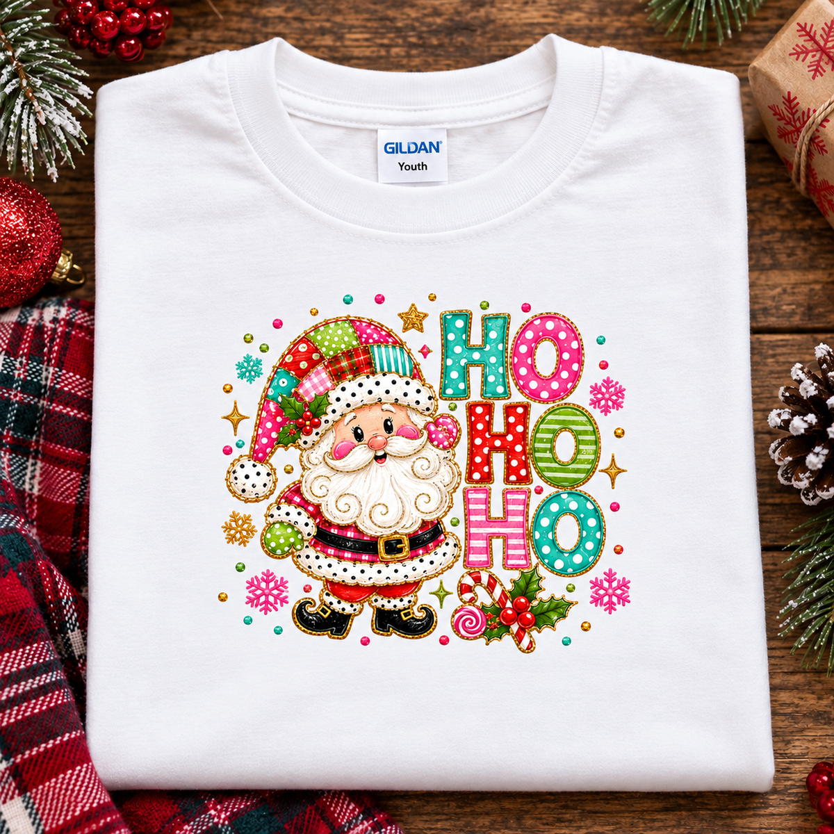 A folded white Gildan Youth t-shirt featuring a colorful Christmas graphic of Santa Claus and the text 'HO HO HO' in patterned letters, styled on a rustic wooden background with holiday decorations.