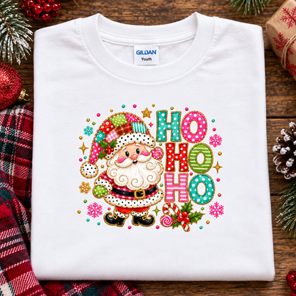 A folded white Gildan Youth t-shirt featuring a colorful Christmas graphic of Santa Claus and the text 'HO HO HO' in patterned letters, styled on a rustic wooden background with holiday decorations.
