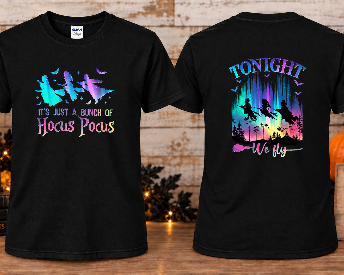 Black short-sleeve T-shirt shown front and back on a wooden surface, decorated with Halloween-themed props including a pumpkin, books, and string lights. The front graphic shows witch silhouettes and the text 'IT'S JUST A BUNCH OF Hocus Pocus' in a pastel rainbow gradient.