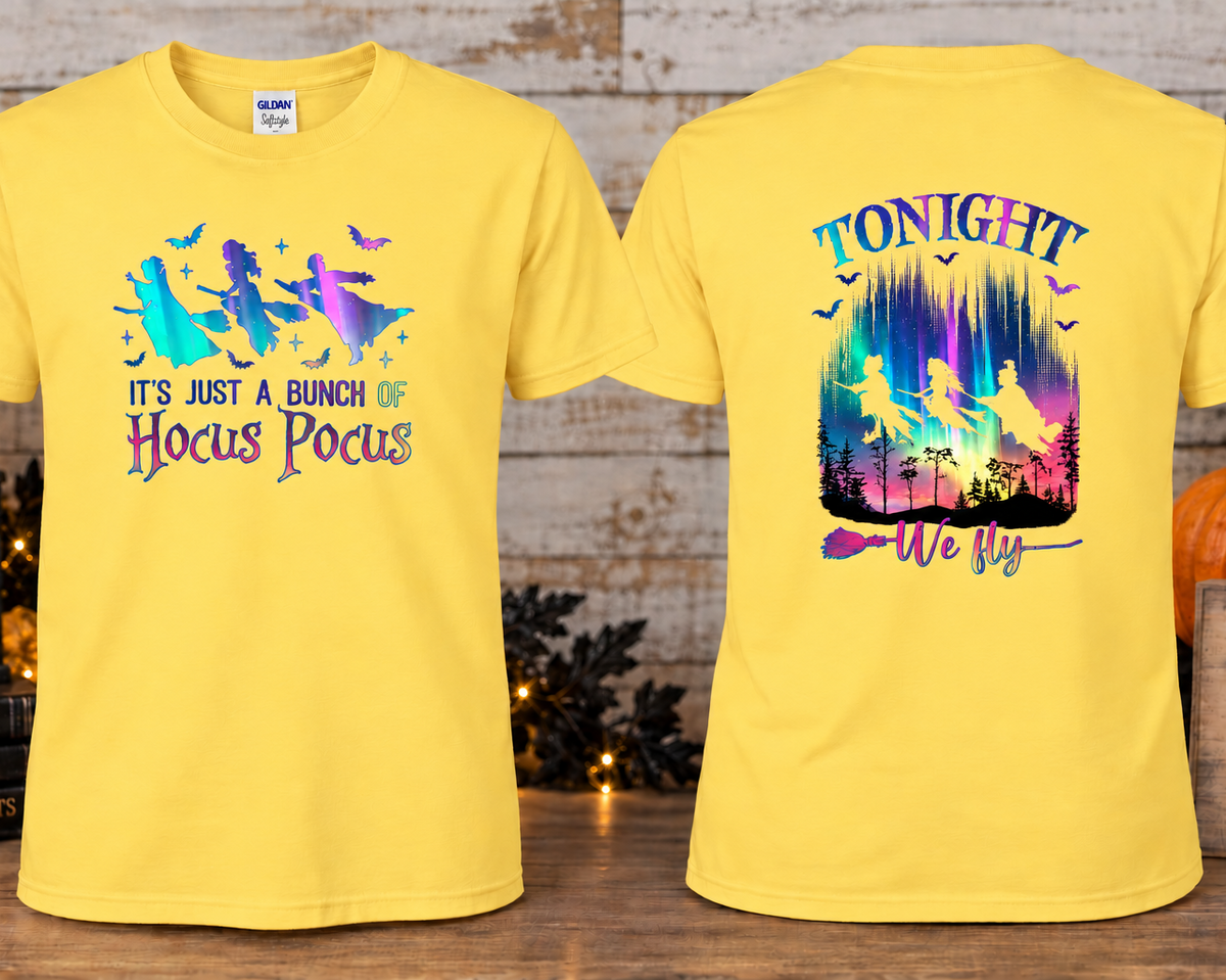 A daisy colored graphic T-shirts shown front and back on a styled wooden surface with autumn props. The front has a colorful witch flying silhouette graphic with the text 'IT'S JUST A BUNCH OF Hocus Pocus.