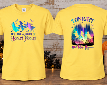 A daisy colored graphic T-shirts shown front and back on a styled wooden surface with autumn props. The front has a colorful witch flying silhouette graphic with the text 'IT'S JUST A BUNCH OF Hocus Pocus.