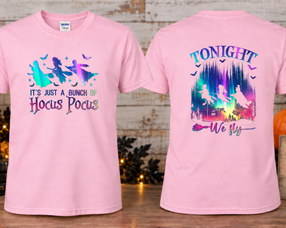 A light pink short-sleeve graphic T-shirts shown front and back on a styled tabletop setup. The front has a witch silhouette graphic with the text 'IT'S JUST A BUNCH OF Hocus Pocus.