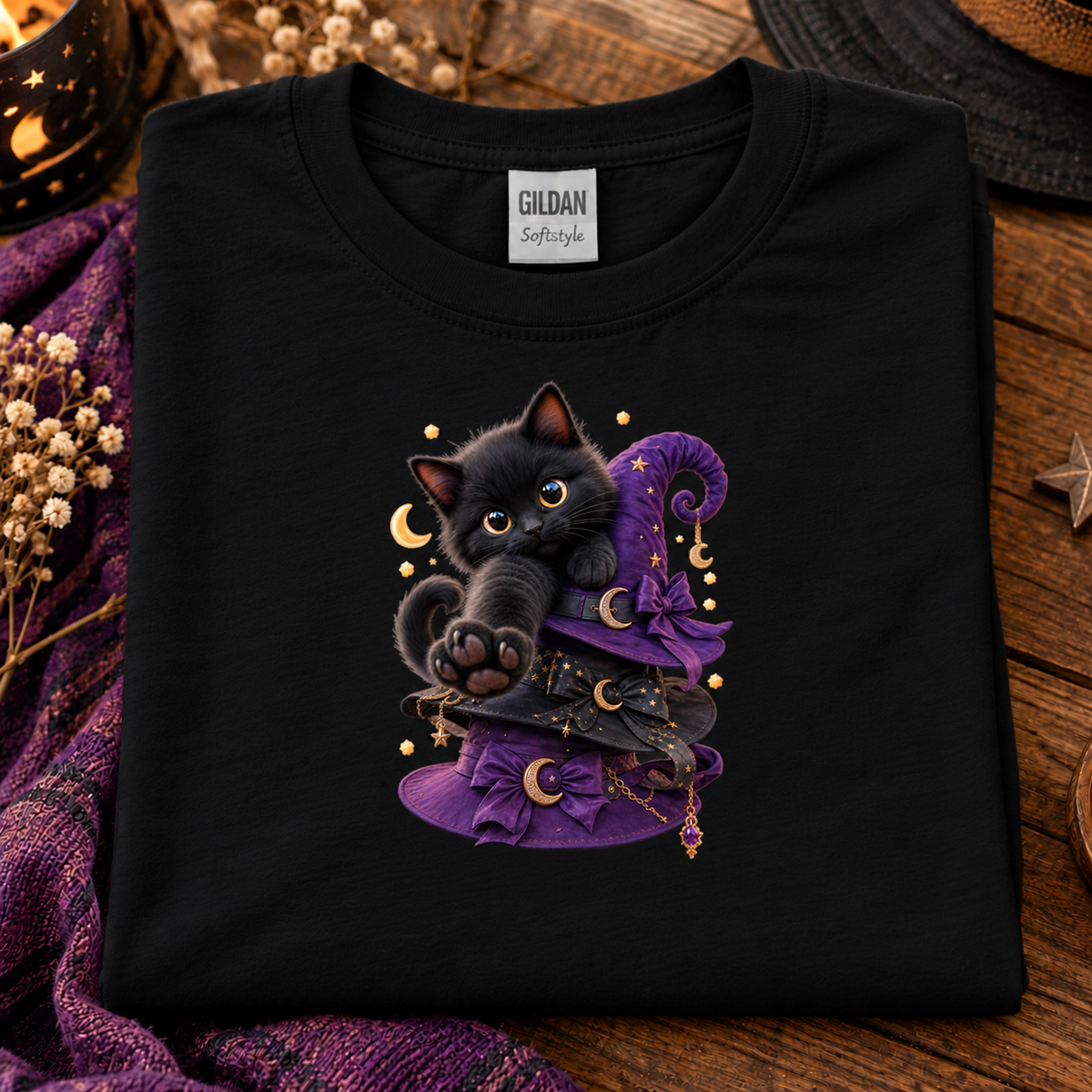 A folded black Gildan Softstyle t-shirt with a graphic of a cute black kitten wearing a purple witch hat, displayed on a rustic wooden surface with dried flowers and a purple blanket.