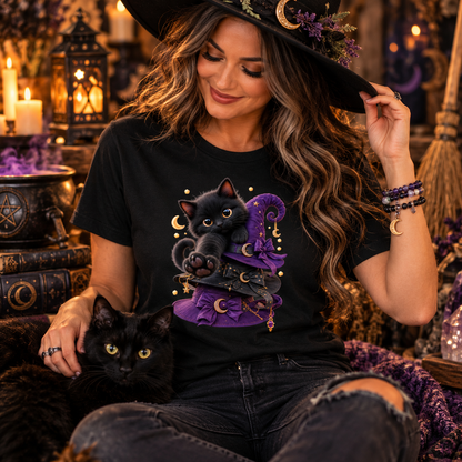 A woman in a witch hat and black distressed jeans sits indoors wearing a black graphic T-shirt with a black cat and purple witch hat illustration. A black cat rests on her lap in a cozy, candle-lit, mystical room.