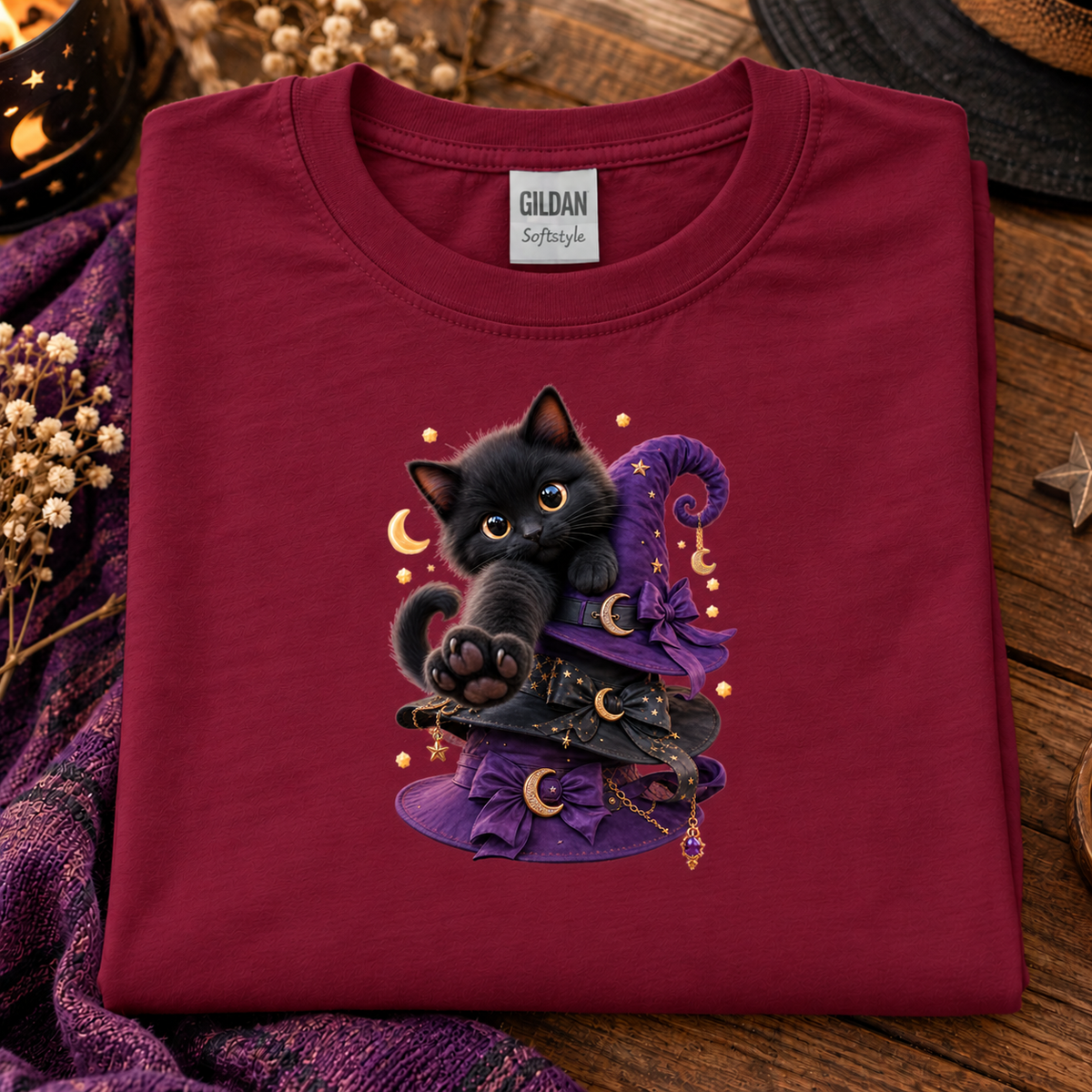 A folded cardinal red t-shirt with a graphic of a cute black kitten sitting on a purple witch hat, displayed on a rustic wooden surface with cozy decorative props.