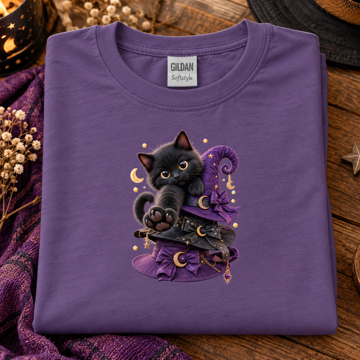 A folded heather purple Gildan Softstyle t-shirt with a graphic of a black kitten wearing a purple witch hat and holding gold crescent moons, styled on a rustic wooden surface with purple fabric and dried flowers.