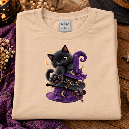 A natural folded t-shirt with a graphic of a black kitten wearing a purple witch hat, displayed on a rustic wooden surface with decorative purple fabric and dried flowers.