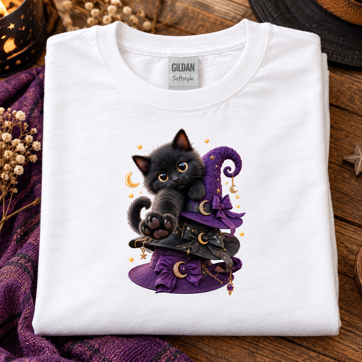 A folded white Gildan Softstyle T-shirt with a cute black kitten illustration wearing a purple witch hat, displayed on a rustic wooden surface with purple fabric and dried flowers.