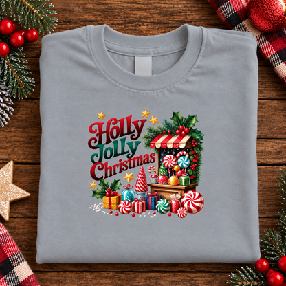 A folded sport gray crewneck t-shirt with a colorful 'Holly Jolly Christmas' graphic print, displayed on a rustic wooden background with holiday decorations like pine branches, red berries, and a plaid blanket.