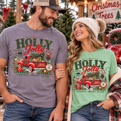 A man and a woman standing together in a festive holiday setting, both wearing matching graphic t-shirts. The man wears a heathered gray t-shirt and the woman wears a sage green t-shirt, both featuring a vintage red truck graphic with Christmas trees and the text 'HOLLY JOLLY WINTER CHRISTMAS'.