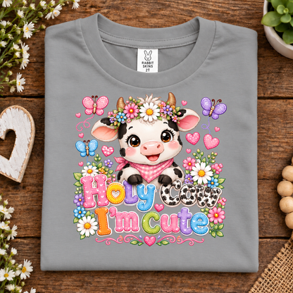 A folded titanium gray t-shirt with a colorful cartoon cow graphic and the text 'Holy cow I'm cute' on a rustic wooden background.