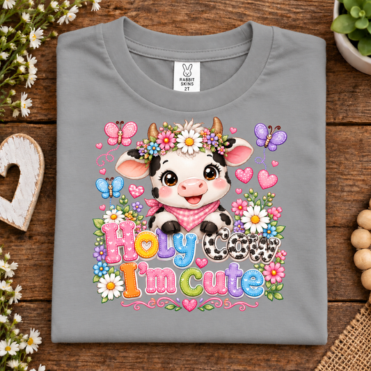 A folded titanium gray t-shirt with a colorful cartoon cow graphic and the text 'Holy cow I'm cute' on a rustic wooden background.