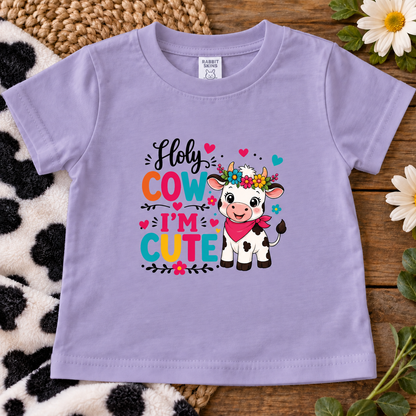 A lavender short-sleeve baby t-shirt with a colorful graphic that reads 'Holy COW I'm CUTE' next to a cartoon cow, laid flat on a rustic wood background with a cow-print blanket and flowers.