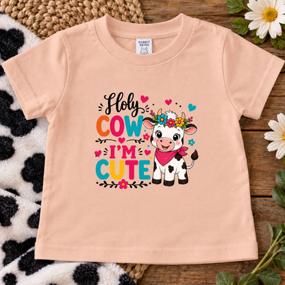A peach-colored short-sleeve baby t-shirt with a colorful graphic of a cute cartoon cow and the text 'Holy COW I'M CUTE'. The t-shirt is displayed flat on a rustic wooden surface with a black-and-white cow print blanket, woven mat, and white flowers.