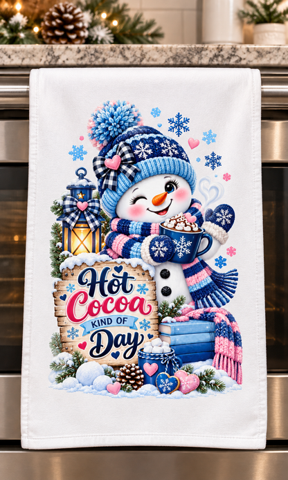 A white kitchen towel with a festive winter design featuring a snowman in a blue plaid hat and scarf, holding a mug of hot cocoa, with the text 'Hot Cocoa KIND OF Day'. The towel is hanging in front of a stainless steel oven.