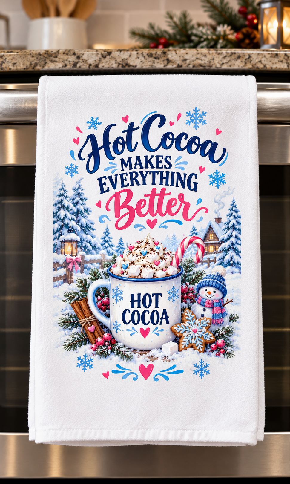 A white kitchen towel hanging on a stainless steel oven handle, featuring a festive Christmas-themed hot cocoa graphic with the text 'Hot Cocoa Makes Everything Better' and an illustration of a mug filled with cocoa, snowflakes, pine trees, candy canes, and a snowman.