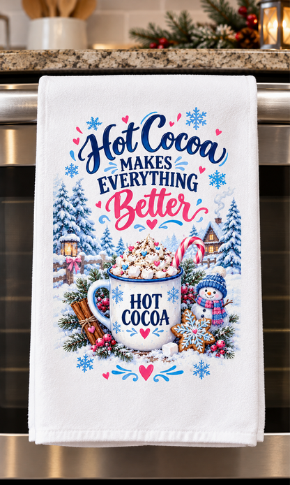 A white kitchen towel hanging on a stainless steel oven handle, featuring a festive Christmas-themed hot cocoa graphic with the text 'Hot Cocoa Makes Everything Better' and an illustration of a mug filled with cocoa, snowflakes, pine trees, candy canes, and a snowman.