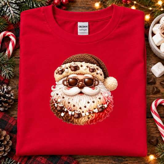A folded red Gildan Signature t-shirt featuring a festive Santa Claus face graphic made of candy canes, marshmallows, and chocolate, displayed on a rustic wooden background with Christmas decorations.