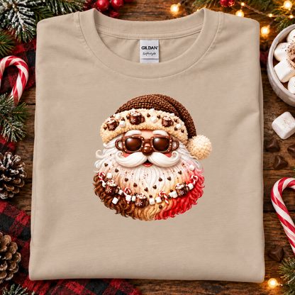 A sand colored Gildan Signature T-shirt laid flat on a rustic wooden surface decorated with Christmas greenery, candy canes, and warm string lights. The shirt features a large graphic of a Santa face made from whipped cream, cocoa powder, marshmallows, and hot chocolate toppings.