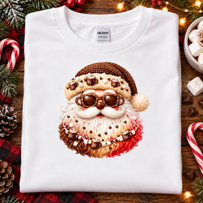 A folded white Gildan Softstyle T-shirt with a large festive Santa face graphic made from candy, marshmallows, and hot chocolate, styled on a wooden surface with holiday decorations.