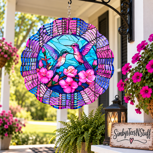A colorful hanging suncatcher or garden ornament featuring two hummingbirds and pink flowers, displayed on a porch with flowers, greenery, and a lantern in the background.