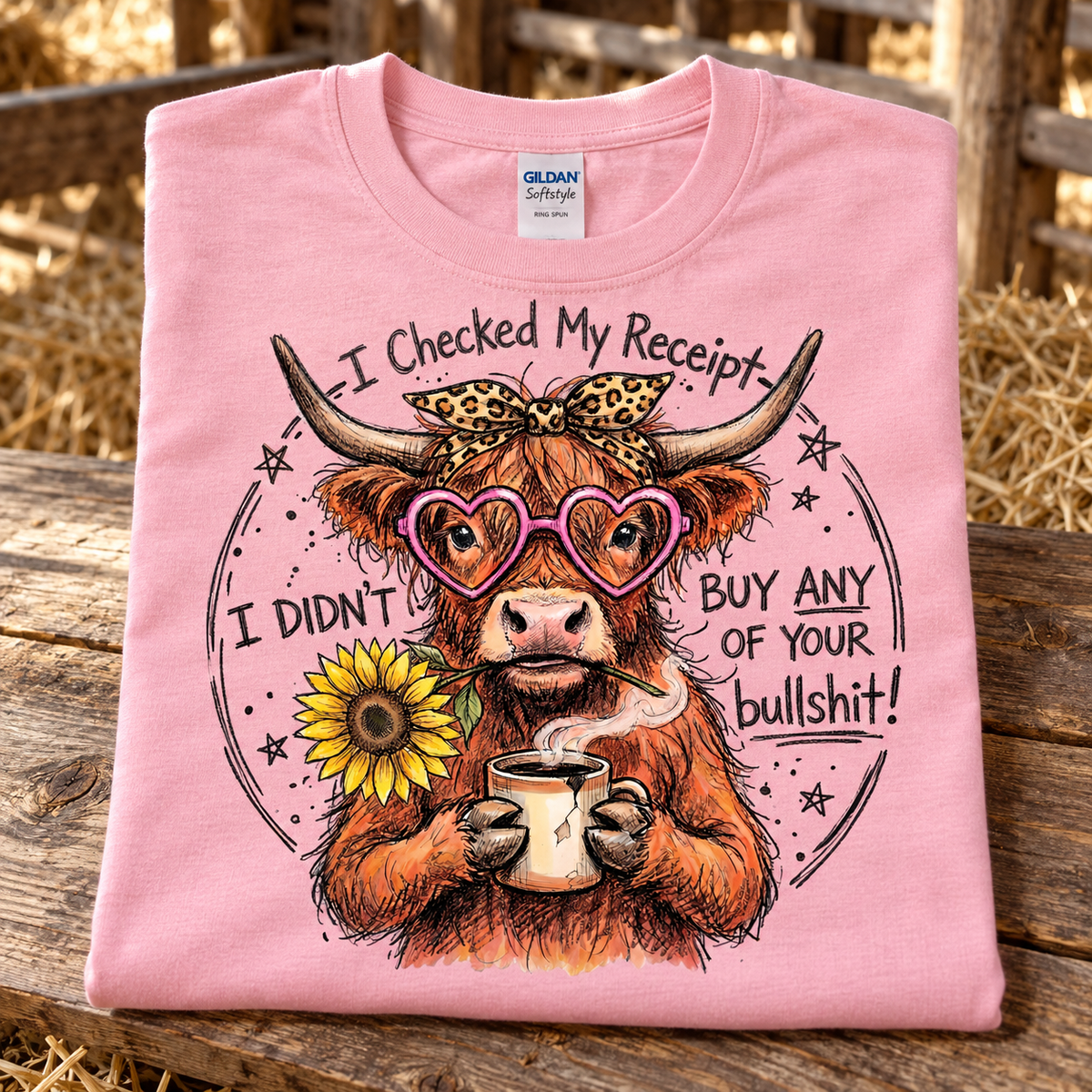 A folded light pink Gildan Softstyle t-shirt is displayed on a rustic wooden surface with a blurred hay bale background. The shirt features a graphic of a highland cow wearing heart-shaped glasses and a leopard print bow, holding a mug with steam, surrounded by the text 'I Checked My Receipt I Didn't Buy Any of Your bullshit!' and a yellow sunflower.