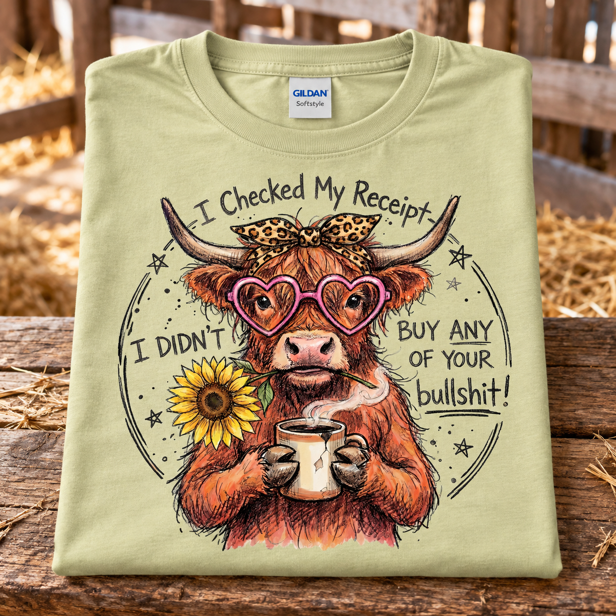 A folded pistachio green Gildan Softstyle t-shirt is displayed on a rustic wooden surface with a blurred barn background. The shirt features a detailed graphic of a Highland cow wearing pink heart-shaped glasses, a leopard-print bow, and holding a sunflower and a mug, with the text 'I Checked My Receipt I DIDN'T BUY ANY OF YOUR bullshit!'.