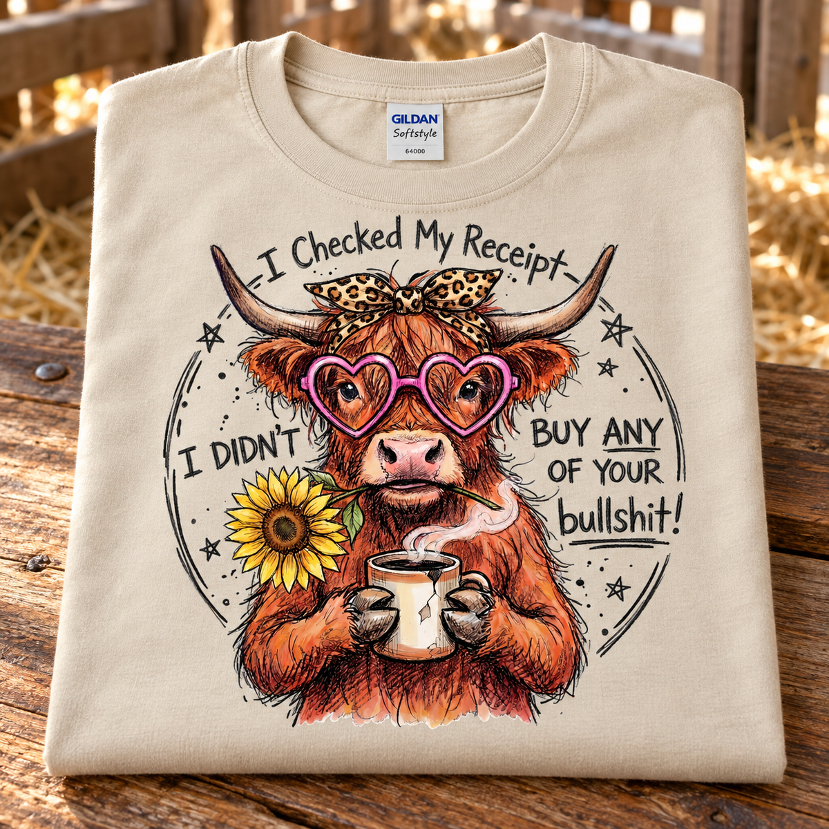 A folded sand Gildan Softstyle t-shirt is displayed on a rustic wooden surface with a blurred outdoor background. The shirt features a graphic of a highland cow wearing pink heart-shaped sunglasses and a leopard print bow, holding a mug with a sunflower in front of it.