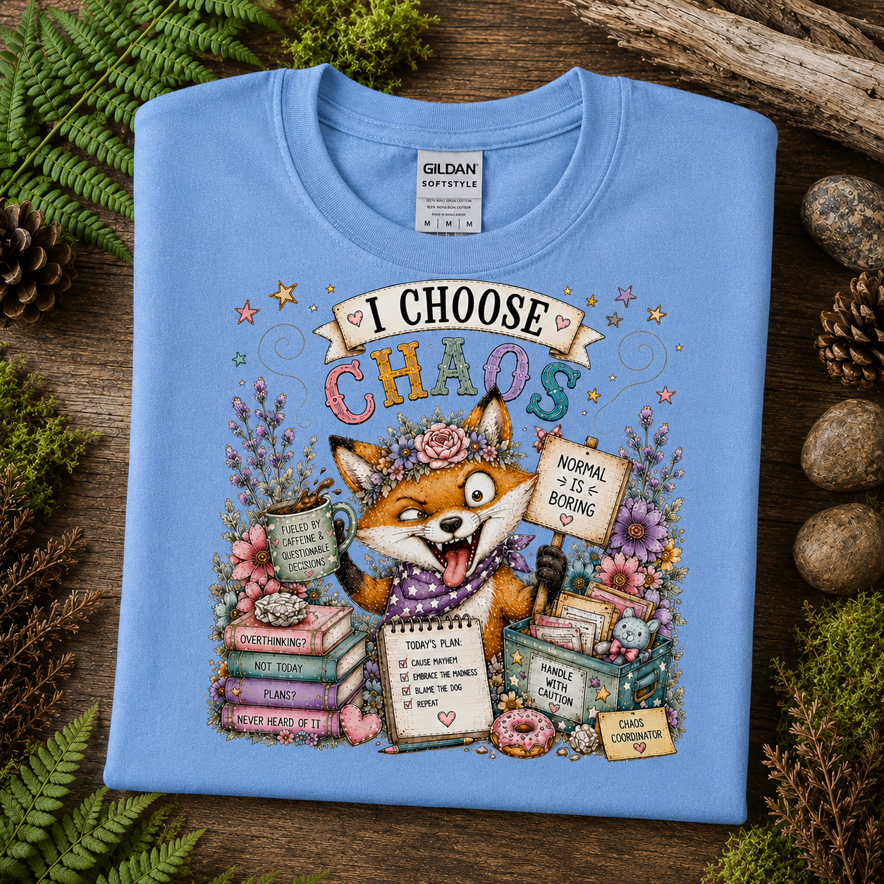 A folded carolina blue Gildan Softstyle T-shirt is displayed on a rustic wooden surface with green ferns, pinecones, and small stones around it. The shirt features a colorful illustrated graphic of a fox surrounded by books, flowers, and signs with the text 'I CHOOSE CHAOS'.