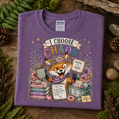 A folded heather purple Gildan Softstyle t-shirt with a colorful cartoon fox graphic and the text 'I CHOOSE CHAOS', displayed on a rustic wooden surface with green ferns and pinecones.