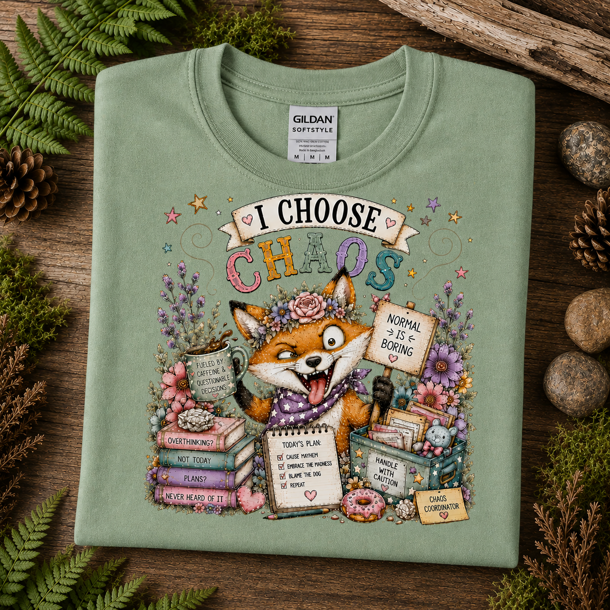 A sage green crewneck t-shirt by Gildan Softstyle, featuring a whimsical graphic of a fox surrounded by books, flowers, and a mug that reads 'I CHOOSE CHAOS'. The shirt is displayed in a flat-lay arrangement on a rustic wooden background with pinecones and ferns.