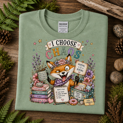 A sage green crewneck t-shirt by Gildan Softstyle, featuring a whimsical graphic of a fox surrounded by books, flowers, and a mug that reads 'I CHOOSE CHAOS'. The shirt is displayed in a flat-lay arrangement on a rustic wooden background with pinecones and ferns.