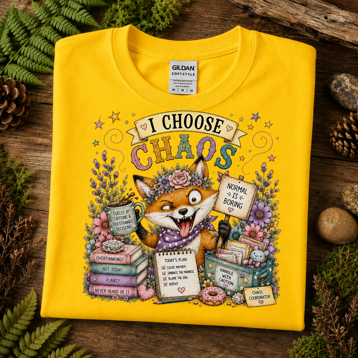 A folded daisy colored Gildan SofteStyle t-shirt is displayed on a rustic wooden surface surrounded by green ferns, pinecones, and dried leaves. The front of the shirt features a colorful graphic of a fox character surrounded by books, flowers, and signs with text like 'I CHOOSE CHAOS' and 'NORMAL IS & BORING'.