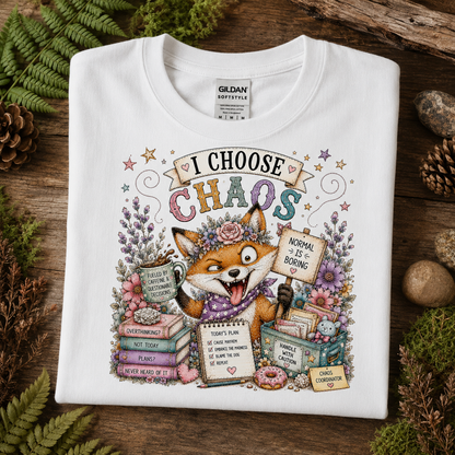 A folded white Gildan Softstyle T-shirt with a colorful illustrated graphic of a fox surrounded by books, flowers, and signs reading 'I CHOOSE CHAOS' and 'NORMAL IS BURNING,' styled on a rustic wood surface with greenery and pinecones.