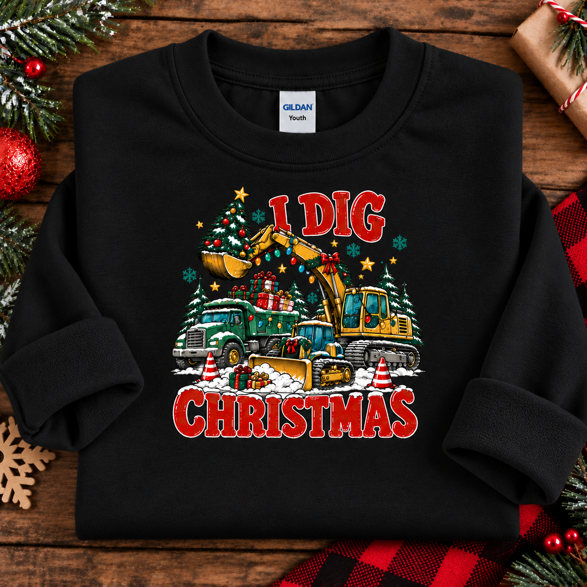 A black long-sleeve crewneck sweatshirt with a festive graphic reading 'I DIG CHRISTMAS' in red letters, featuring construction vehicles and a Christmas tree. The sweatshirt is displayed flat on a rustic wooden background decorated with holiday ornaments, pine branches, and a plaid blanket.