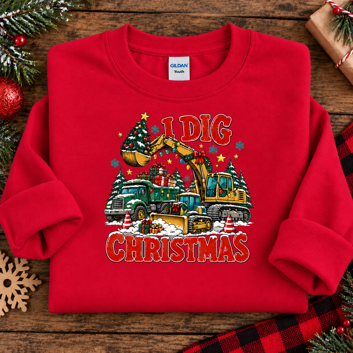 A folded red crewneck sweatshirt on a rustic wooden background with holiday decorations. The sweatshirt features a graphic print of construction vehicles, Christmas trees, snow, and the words 'ELD CHRISTMAS' in red.