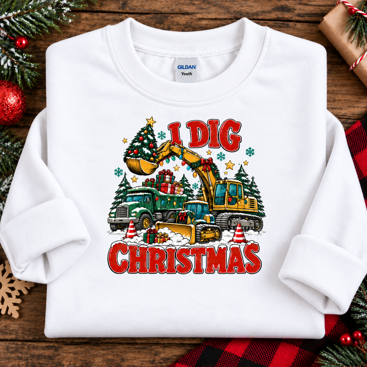 A white Gildan Youth crewneck sweatshirt laid flat on a rustic wooden surface, decorated with a colorful graphic that reads 'I DIG CHRISTMAS' with construction vehicles and Christmas trees. The sweatshirt is styled with holiday decorations like pine branches, ornaments, and a red plaid fabric.