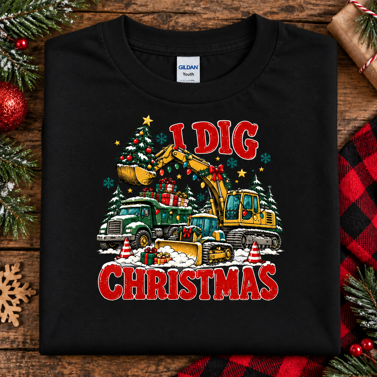 A folded black Gildan Youth t-shirt with a colorful Christmas construction graphic reading 'I DIG CHRISTMAS' is displayed on a rustic wooden background with holiday decorations.