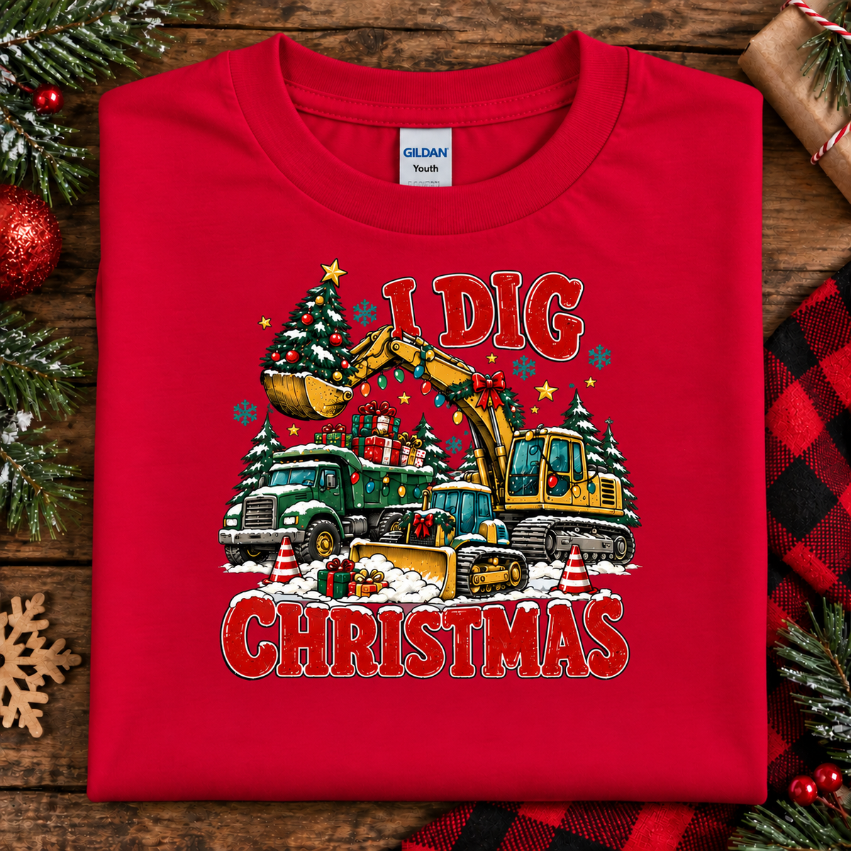 A folded red Gildan Youth t-shirt with a festive 'I Dig Christmas' graphic, displayed on a rustic wooden background with holiday decorations like pine branches, a red ornament, and a plaid blanket.
