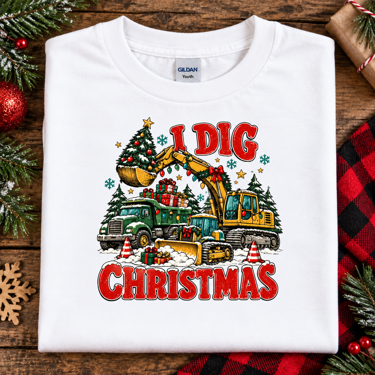 A folded white Gildan Youth t-shirt with a colorful 'I Dig Christmas' graphic featuring construction vehicles and a Christmas tree, displayed on a rustic wooden background with holiday decorations.